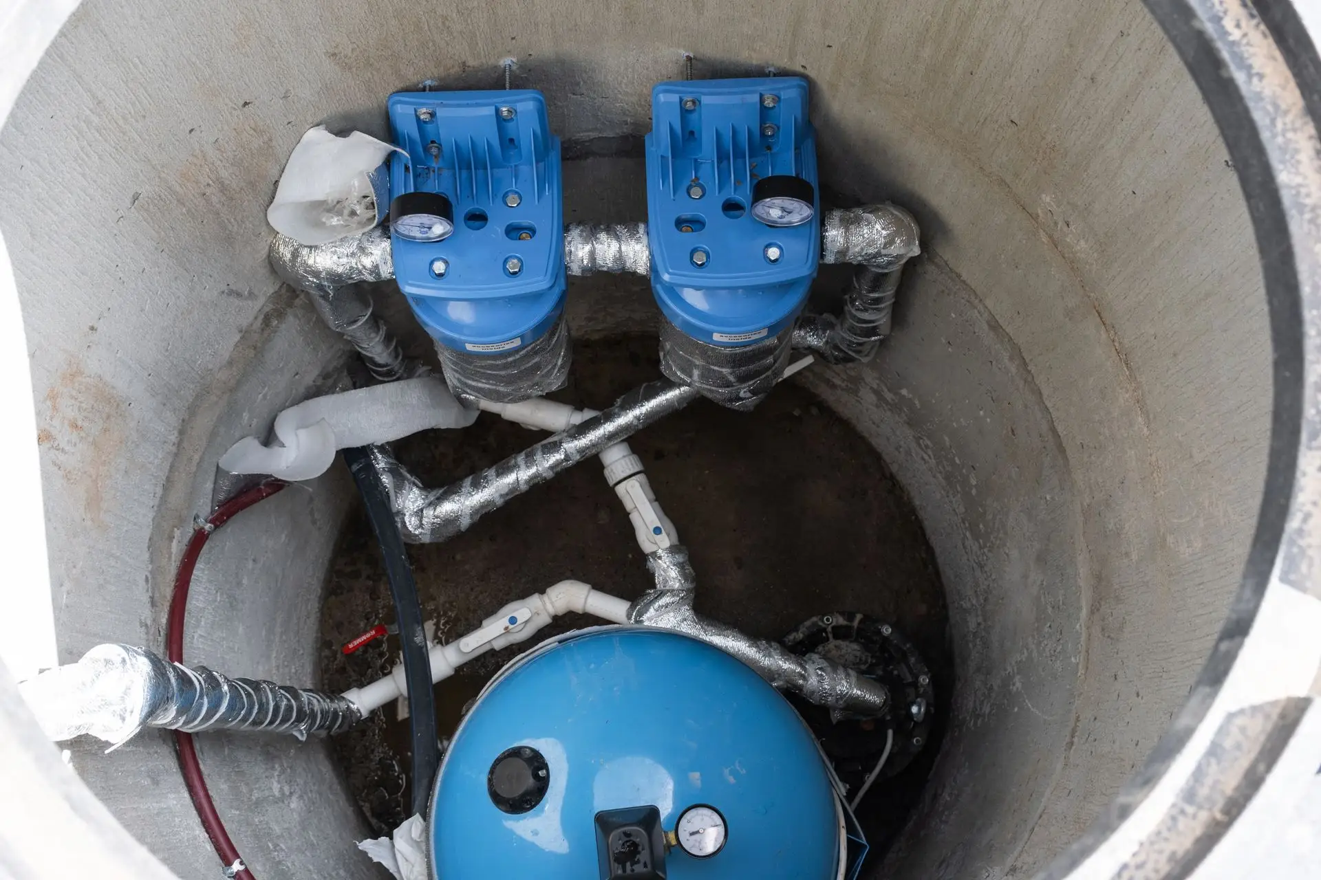 Blue pressure switches, insulated piping, and a pressure tank inside a concrete well pit.