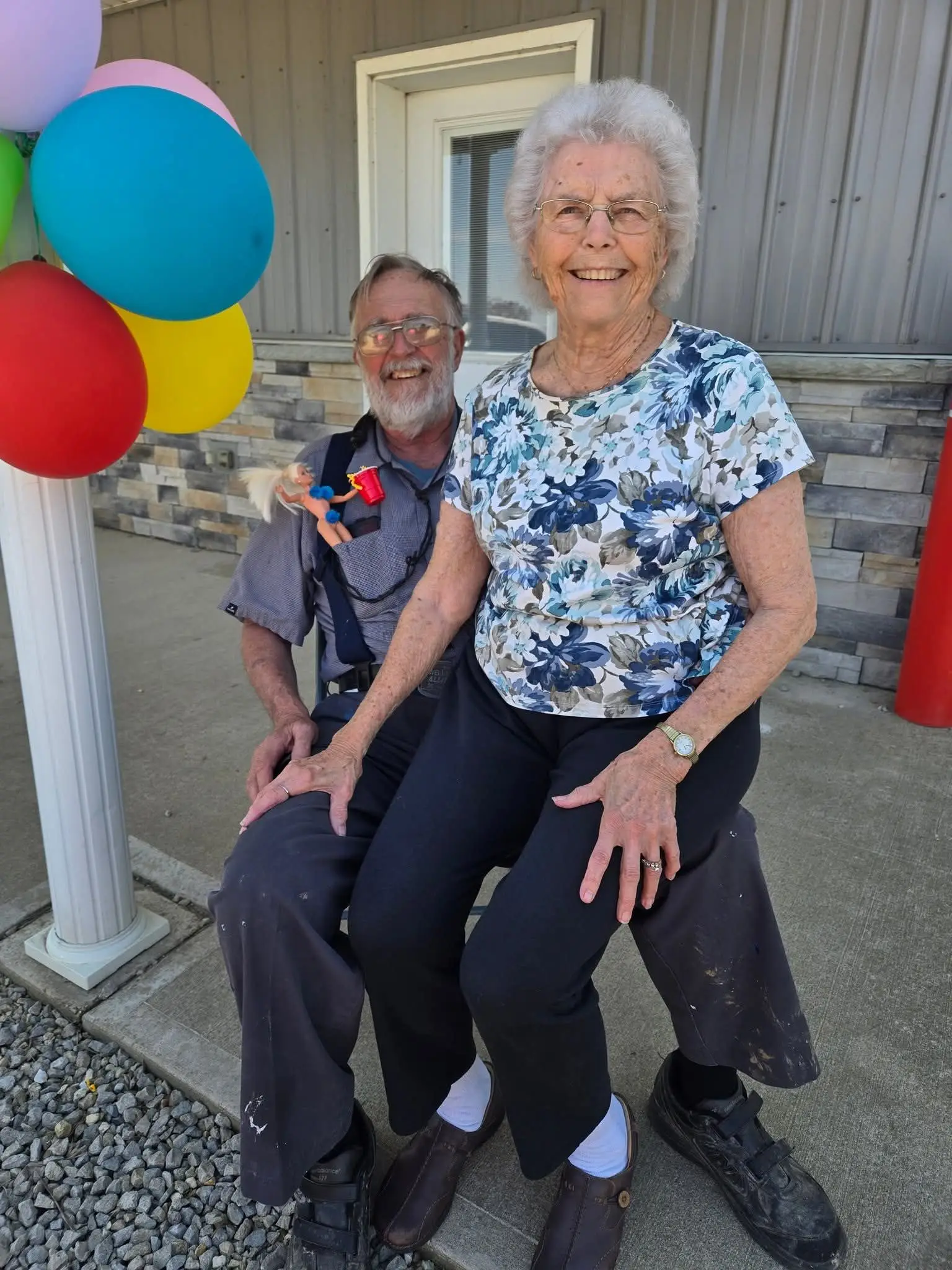 A person in a patterned top sits on the lap of another person in a gray shirt, both smiling next to colorful balloons.