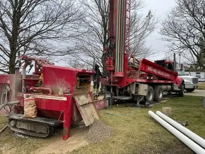 A red rotary drilling rig and mud pit set up in a residential yard, with white casing pipe on the grass